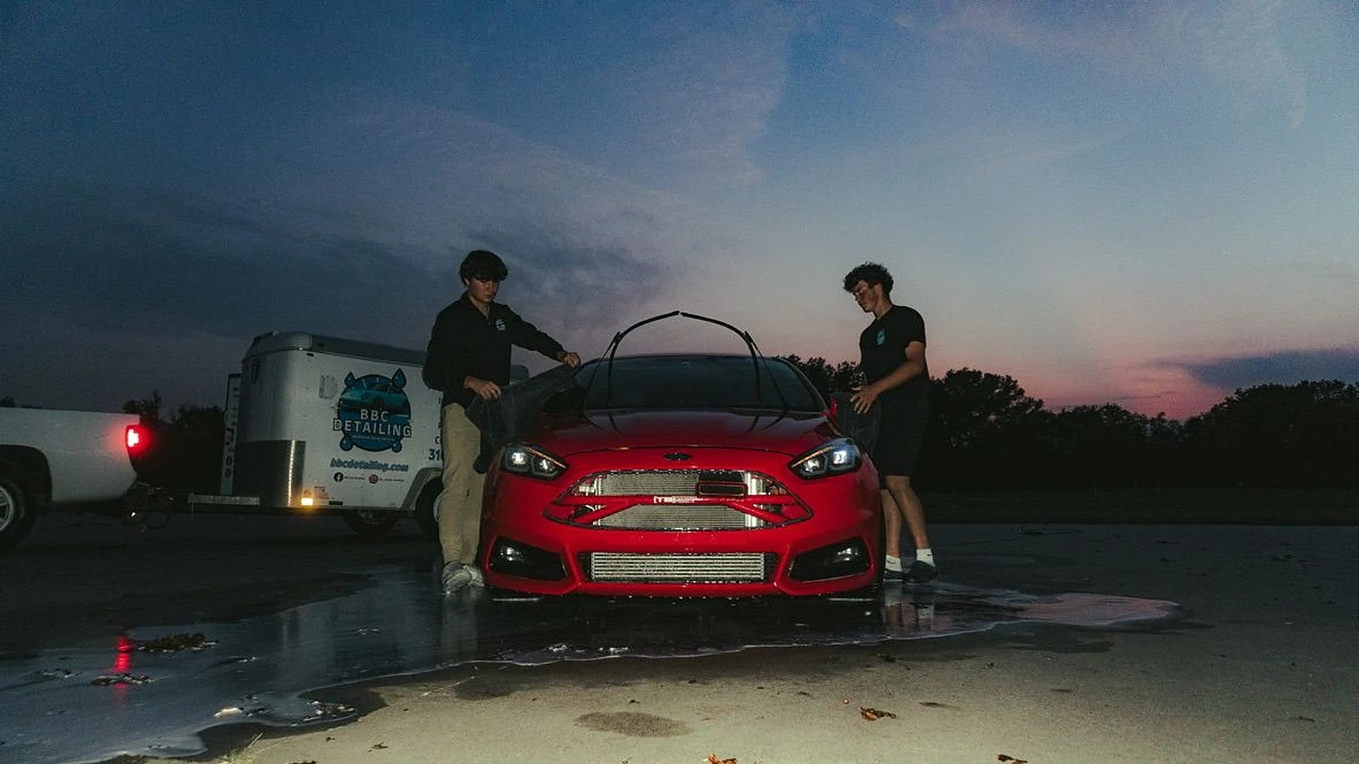 The Blue Badge Customs crew detailing a red Ford Focus ST at dusk beside the shop trailer